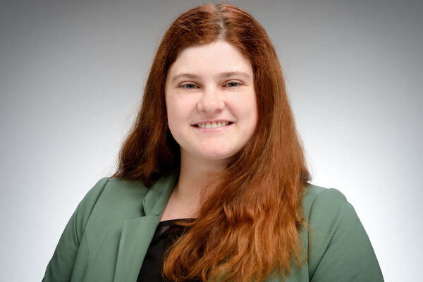 A smiling woman with long reddish-brown hair and a dark green blazer looks forward against a gray background.